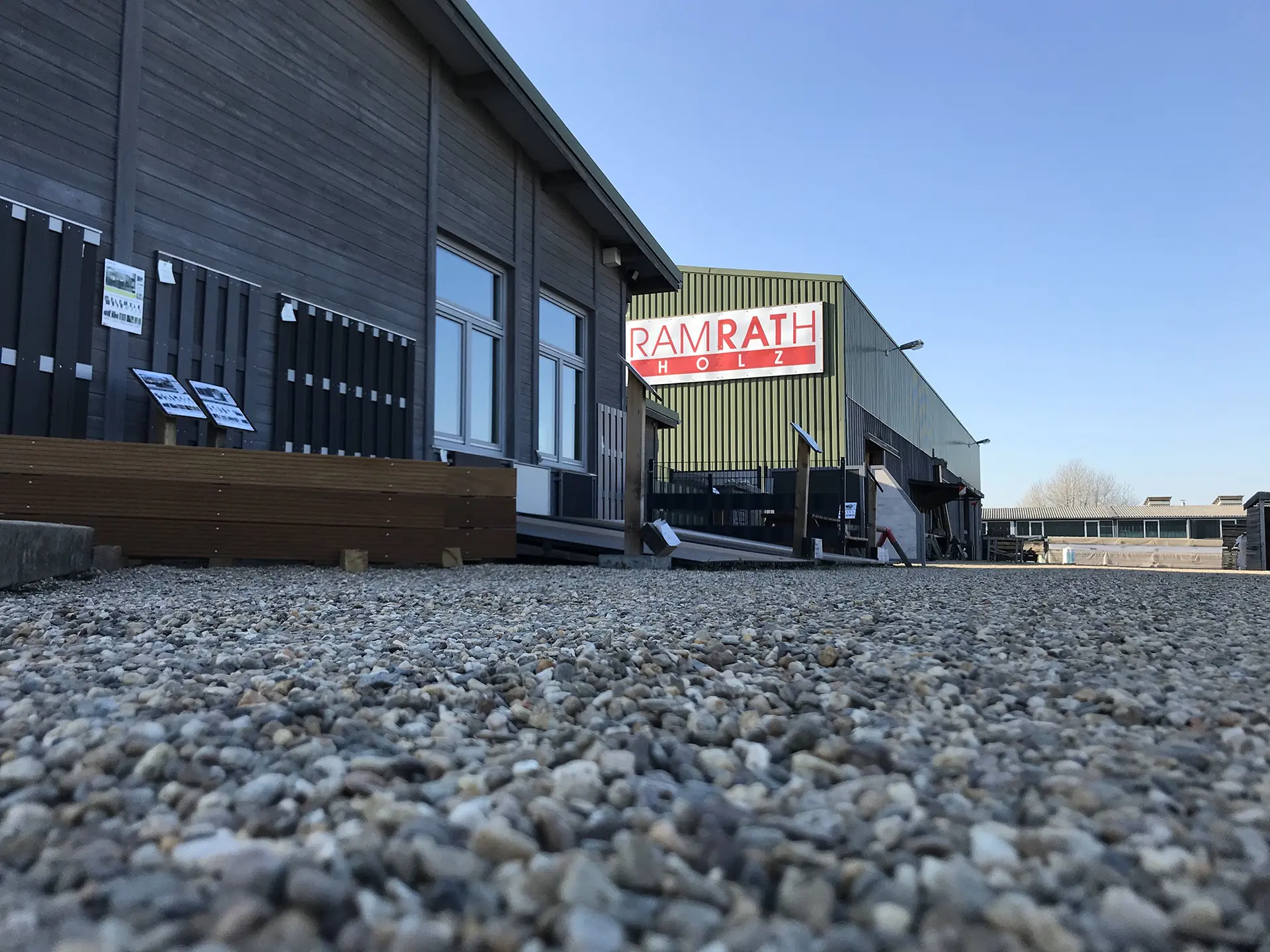 Ausstellungsgelände mit Zäunen, Terrassen und Lagerhalle – Blick von der Einfahrt haus und garten sortiment - startseite - Einfahrt mit Blick auf die Ausstellung für Zäune und Terrassen sowie die Lagerhalle im Hintergrund.